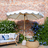 Lifestyle image of Blue Holly- Handcrafted frame made in the UK. Beautiful plain natural white parasol with blue scallop edged. Simple, chic and elegant. FSC certified ash frame made in Hampshire with our own designs of brass fittings and pulleys. A parasol perfect for a picnic, patio, through your table with an umbrella hole or by your sun lounger at the pool.
