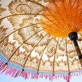 Helena Round Bamboo Parasol - soft beige twill with lotus design hand-painted in gold ink. Inside the threading is vibrant tangerine orange, and the fringe is a combination of peony pink, black and orange tassels.
